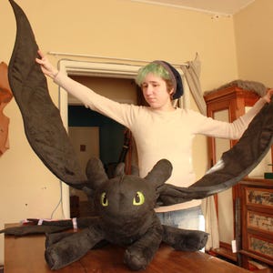May include: A large, plush black dragon toy with large wings and yellow eyes. The dragon is lying on a wooden surface. The wings are spread wide. The dragon's wingspan appears to be approximately 60 inches.