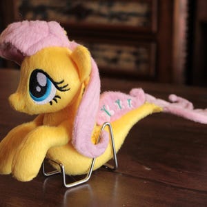 Puede incluir: Un peluche de pony amarillo con melena y cola rosa, y ojos azules. El juguete está tumbado sobre un soporte metálico. El pony tiene un tocado rosa y una cola rosa con tres acentos azul claro.
