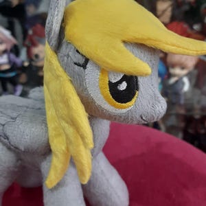 Puede incluir: Un peluche de pony con pelaje gris y pelo amarillo. El pony tiene una cara bordada con un gran ojo amarillo y una pupila negra. El juguete está hecho de tela suave y tiene una melena y cola cosidas.
