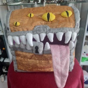 Puede incluir: Un cofre de peluche con diseño de monstruo. El cofre es marrón con ribetes grises, dientes blancos y ojos amarillos. Una lengua rosa se extiende desde la boca. Tiene un ojo de cerradura y una cerradura.