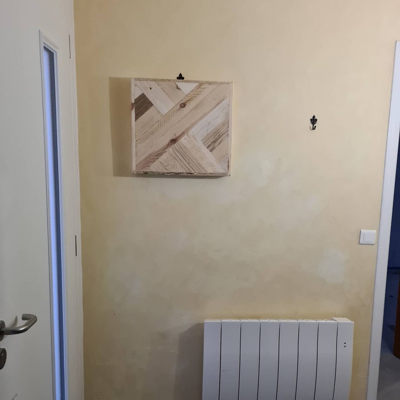 May include: A square wooden wall art piece with a geometric design made of light-coloured wood planks. The artwork is mounted on a wall with a light yellow paint. A black hook is above the artwork.