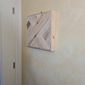 May include: A square wooden wall art piece with a geometric pattern. The artwork is made of light-coloured wood planks arranged in a diagonal design. The piece is mounted on a wall with a light yellow colour.