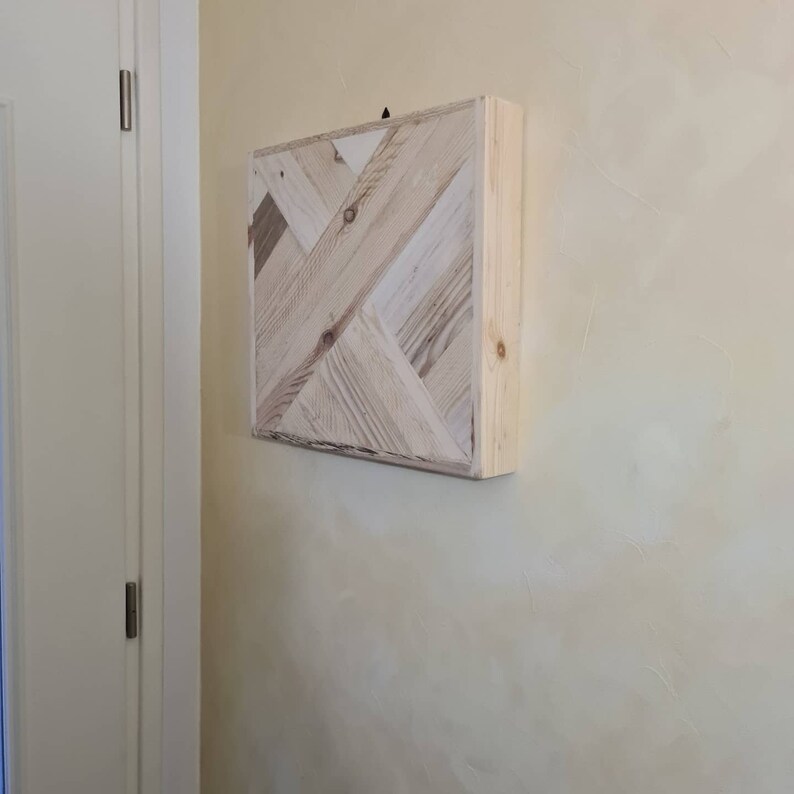 May include: A square wooden wall art piece with a geometric pattern. The artwork is made of light-coloured wood planks arranged in a diagonal design. The piece is mounted on a wall with a light yellow colour.