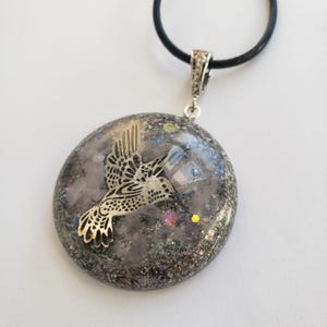 May include: A silver hummingbird pendant with a clear resin background filled with iridescent glitter and small crystals. The pendant is attached to a black cord.