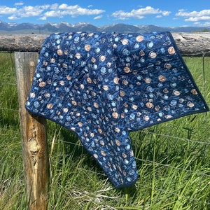 May include: A blue and brown quilted blanket with a pattern of planets and stars. The blanket is draped over a wooden fence post against a backdrop of a blue sky and green grass.