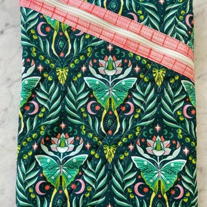 May include: A quilted, rectangular pouch with a dark teal background featuring a repeating pattern of green moths, leaves, and crescent moons. A diagonal, coral-colored zipper with a white zipper pull and a white zipper runs across the top.