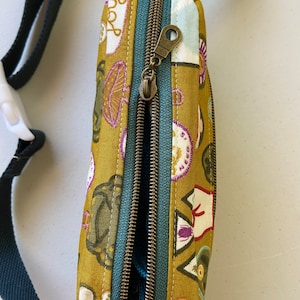 May include: A mustard yellow fabric fanny pack with a teal zipper and a brown zipper pull. The fabric has a pattern of various illustrations including mushrooms, a cat, and a heart with the word "love" on it.