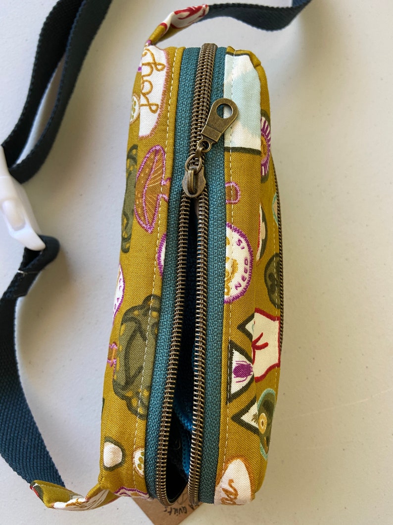 May include: A mustard yellow fabric fanny pack with a teal zipper and a brown zipper pull. The fabric has a pattern of various illustrations including mushrooms, a cat, and a heart with the word "love" on it.