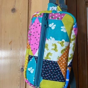May include: A colorful patchwork backpack with a teal zipper and strap. The bag features various patterned fabric sections, including pink stars, yellow polka dots, and tiger stripes. The bag is hanging on a wooden door.