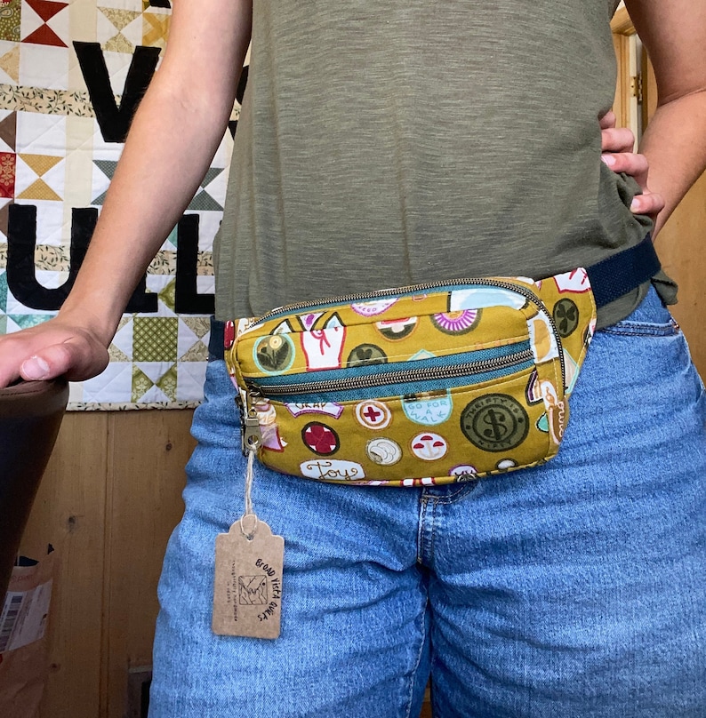 May include: A yellow fabric fanny pack with a variety of colorful patches and the word "Toy" on it. The fanny pack is worn around the waist of a person wearing blue jeans.