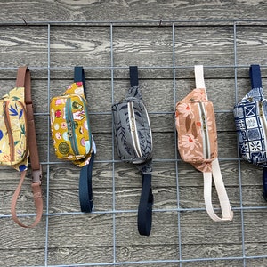 May include: Five different patterned fanny packs hanging on a wire rack. The fanny packs are in various colors and patterns, including yellow, green, brown, and blue.