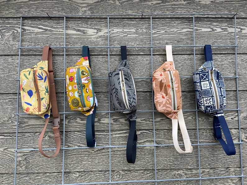 May include: Five different patterned fanny packs hanging on a wire rack. The fanny packs are in various colors and patterns, including yellow, green, brown, and blue.