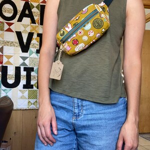 May include: A mustard yellow fanny pack with a black strap and a colorful print of various icons. The pack is worn by a person wearing a green tank top and blue denim shorts.