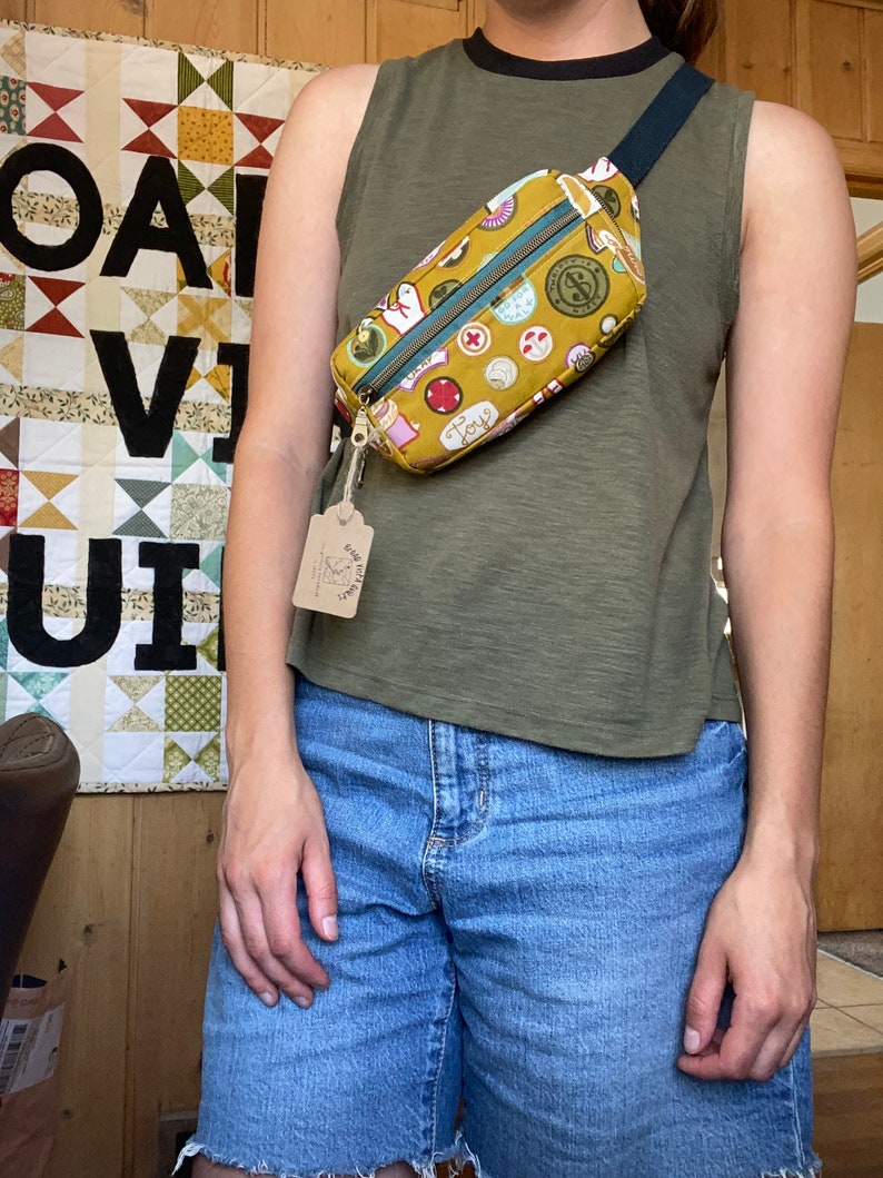 May include: A mustard yellow fanny pack with a black strap and a colorful print of various icons. The pack is worn by a person wearing a green tank top and blue denim shorts.
