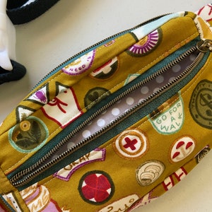 May include: A small mustard yellow fabric fanny pack with a green zipper and a white polka dot lining. The fabric is printed with various colorful patches and text, including "Go for a walk", "Love", and "Need is".