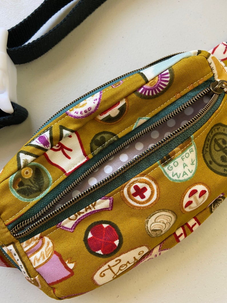 May include: A small mustard yellow fabric fanny pack with a green zipper and a white polka dot lining. The fabric is printed with various colorful patches and text, including "Go for a walk", "Love", and "Need is".