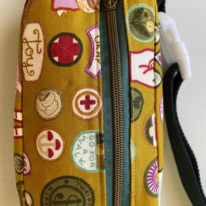 May include: A mustard yellow fabric fanny pack with a zipper closure and a black adjustable strap. The fabric is covered in a pattern of colorful patches with various designs and text, including "Joy", "Go for All", and a red cross.
