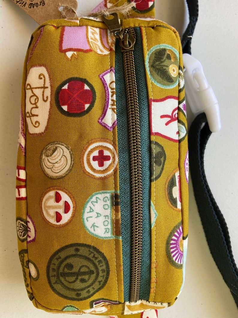 May include: A mustard yellow fabric fanny pack with a zipper closure and a black adjustable strap. The fabric is covered in a pattern of colorful patches with various designs and text, including "Joy", "Go for All", and a red cross.