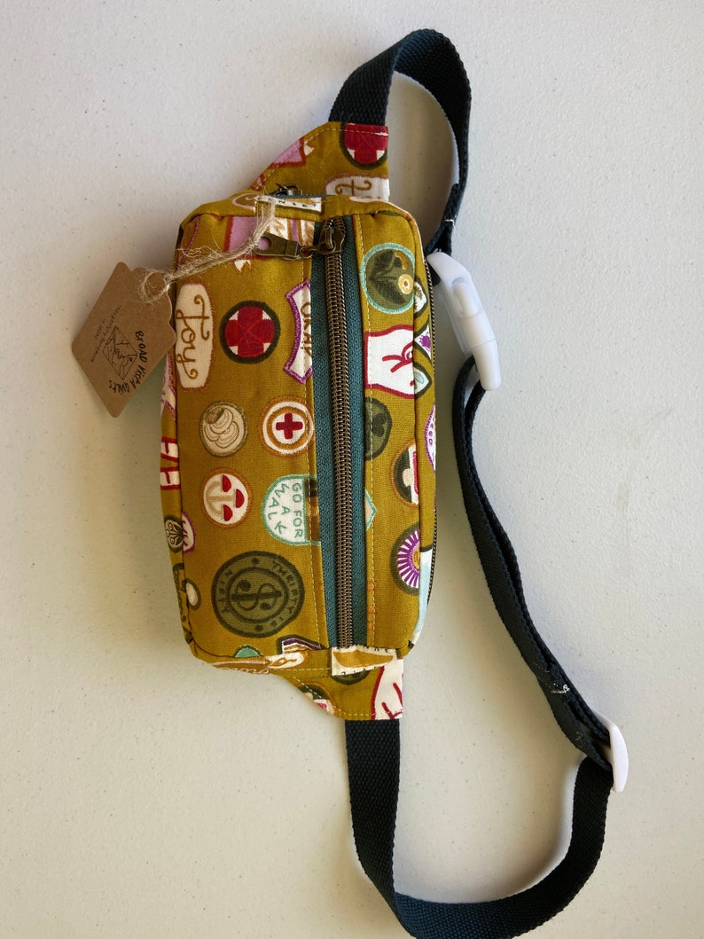 May include: A mustard yellow fanny pack with a black adjustable strap and a zipper closure. The pack is covered in colorful patches with various designs, including a red cross, a heart, and the words "Go for a walk".