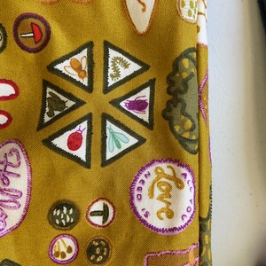 May include: A mustard yellow fabric bag with a variety of colorful patches and designs. The patches include a mushroom, a ladybug, a bee, a caterpillar, a butterfly, a flower, and a speech bubble that says "Hello". The bag also has a patch that says "All you need is love".