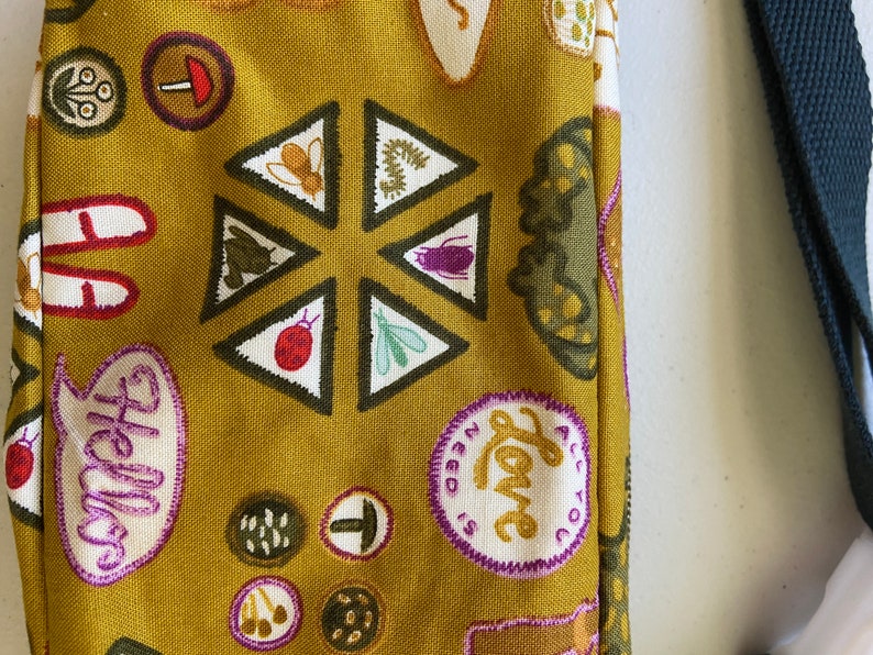 May include: A mustard yellow fabric bag with a variety of colorful patches and designs. The patches include a mushroom, a ladybug, a bee, a caterpillar, a butterfly, a flower, and a speech bubble that says "Hello". The bag also has a patch that says "All you need is love".