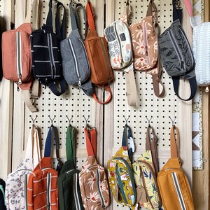 May include: A wall display of 12 colorful fabric fanny packs with various patterns and designs. The fanny packs are hanging on a pegboard with hooks. The fanny packs are made of different fabrics, including floral, geometric, and abstract patterns.