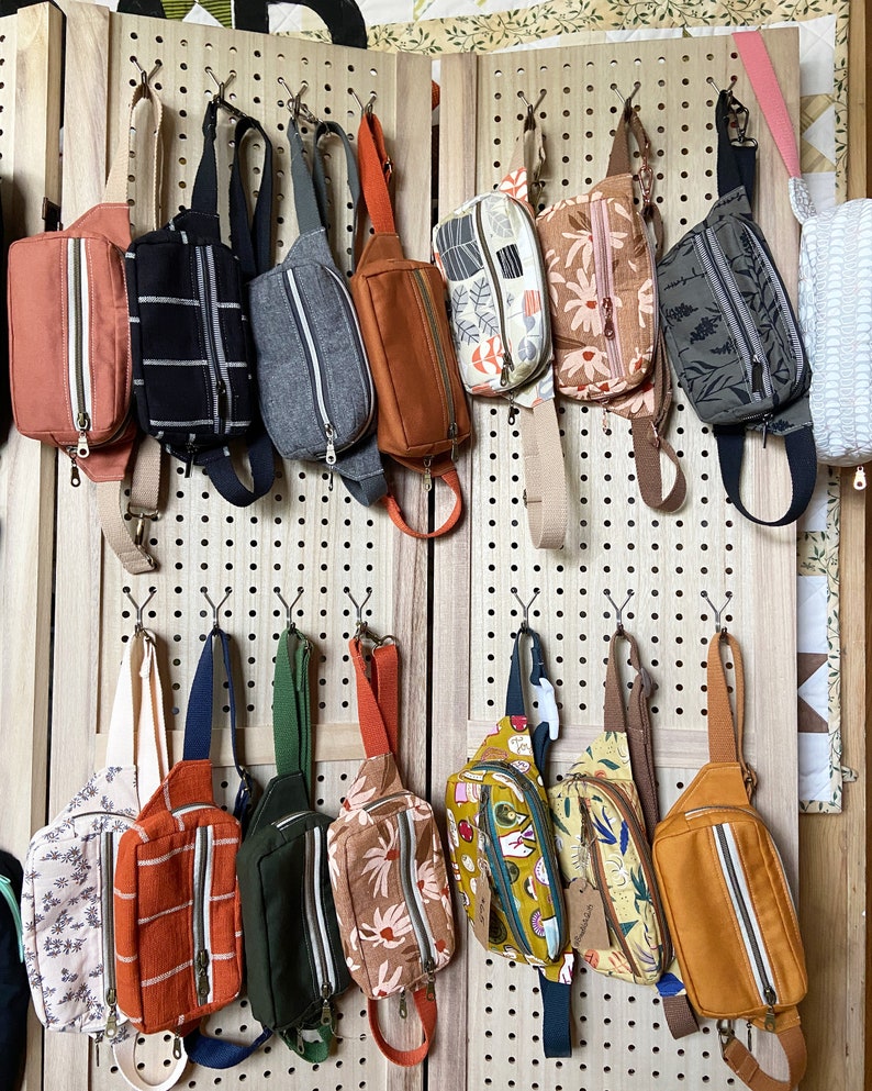 May include: A wall display of 12 colorful fabric fanny packs with various patterns and designs. The fanny packs are hanging on a pegboard with hooks. The fanny packs are made of different fabrics, including floral, geometric, and abstract patterns.
