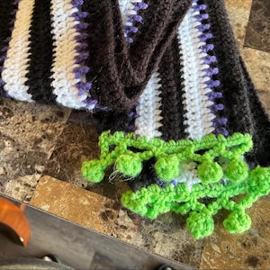 Pode incluir: Uma echarpe de crochê listrada em preto, branco e roxo com pompons verdes nas pontas.