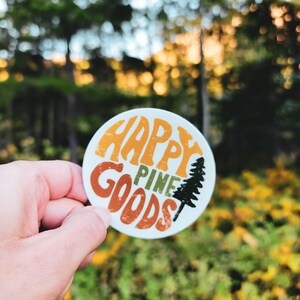 以下が含まれることがあります： オレンジ、緑、茶色で「HAPPY PINE GOODS」の文字が入った丸いステッカー。右側には白黒の松の木のグラフィックがあります。ステッカーは、木や葉のぼやけた背景の前に保持されています。