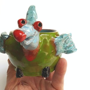 Peut inclure: Un pot de fleurs en céramique fantaisiste en forme d'oiseau. Le pot a un corps vert, des ailes bleues et une tête bleue avec un bec et des yeux rouges. La poterie faite à la main a une finition brillante.