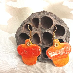 Peut inclure: Une paire de boucles d'oreilles en céramique orange et jaune faites à la main, avec des crochets dorés. Les boucles d'oreilles ont une forme abstraite unique et sont présentées sur un fond neutre. Les boucles d'oreilles mesurent environ 2.5 cm de long.