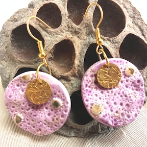Peut inclure: Boucles d'oreilles artisanales avec des pendentifs ronds en céramique lavande texturés. Chaque boucle d'oreille comporte un petit charme doré et des crochets d'oreilles dorés. Les boucles d'oreilles sont présentées sur un fond neutre. Diamètre : environ 3 cm.