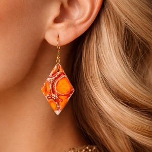 Peut inclure: Boucle d'oreille unique en or avec un pendentif en forme de losange. Le pendentif présente un motif orange et rouge avec des accents blancs. La boucle d'oreille est suspendue à un crochet doré, portée par une personne aux cheveux blonds.
