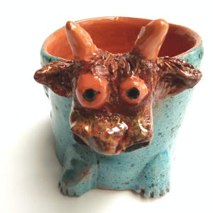 Peut inclure: Pot de fleurs en céramique fait à la main en forme de créature fantaisiste. Le pot a un extérieur bleu clair, un visage brun rougeâtre avec de grands yeux orange et des cornes orange. L'intérieur est orange.