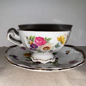 Puede incluir: Juego de taza y platillo blancos con diseño floral, con rosas rosas, flores amarillas y moradas, y ribetes dorados. La taza está llena de un líquido oscuro, posiblemente té o café.