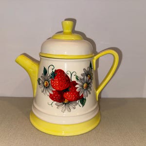 Puede incluir: Tetera de cerámica blanca y amarilla. La tetera tiene una tapa, un asa y un pico. La parte delantera está decorada con fresas rojas y margaritas blancas. Ideal para servir té.