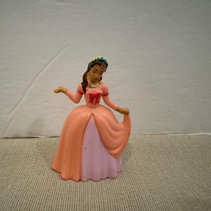 May include: A small plastic princess figurine in a peach-colored gown with a pink underskirt. The princess has brown hair, a blue crown, and is holding her skirt. The figurine is on a neutral-colored surface.