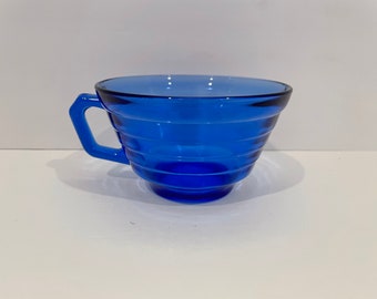 May include: A cobalt blue glass teacup with a flared rim and a handle. The cup has a ribbed design.