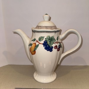 May include: White ceramic teapot with a lid, spout, and handle. The teapot is decorated with a fruit design featuring a pear, grapes, and cherries. The teapot has a base and a decorative rim. A ruler is at the bottom of the image.