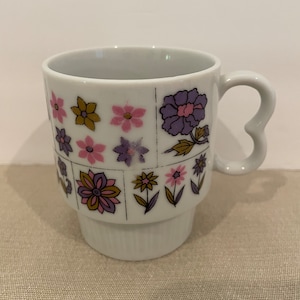 May include: White ceramic mug with a floral pattern in shades of pink, purple, and gold. The mug has a heart-shaped handle.