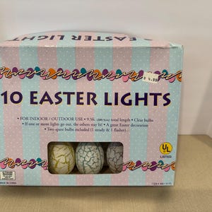 Puede incluir: Caja de 10 luces de Pascua con bombillas en forma de huevo. La caja es de color azul claro con rayas rosas y el texto "10 EASTER LIGHTS". Las luces son para uso en interiores/exteriores y tienen una longitud total de 289,5 cm.