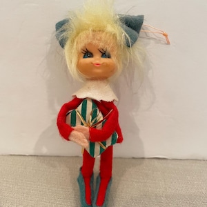 May include: A vintage felt Christmas elf ornament with blonde hair, blue eyes, and a red and white outfit. The elf is holding a green and white striped gift box with a gold ribbon.