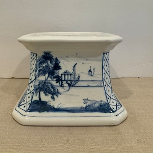 May include: A white ceramic pedestal with a blue and white hand-painted design. The design features a landscape with trees, a building, and a cow. The pedestal has a square top and a square base.