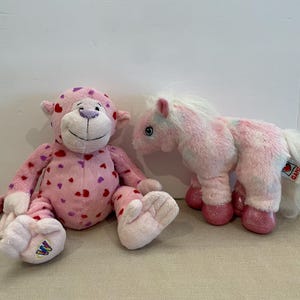 May include: Two pink plush toys, a monkey and a horse, both covered in pink and red hearts. The monkey has a purple nose and the horse has a white mane and tail.