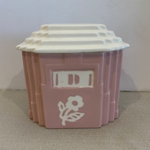 May include: A pink ceramic sugar bowl with a white lid and a white floral design. The bowl has a geometric design on the sides.