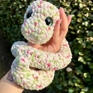 May include: A handmade crochet snake toy in white with pink and green accents. The snake has large, shiny green eyes and is held in a hand. The plush toy is coiled and appears soft and cuddly.
