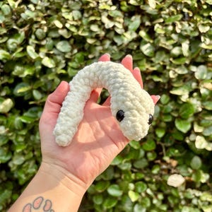 May include: A handmade, off-white crochet snake toy with black safety eyes. The plush snake is held in a hand against a backdrop of green foliage. The snake is made of soft, textured yarn.