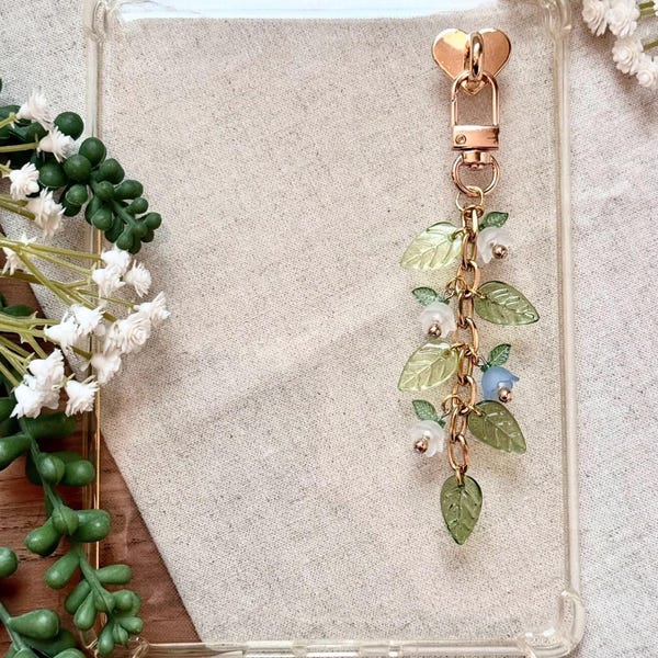 Puede incluir: Funda de teléfono transparente con un cierre dorado en forma de corazón y una cadena que sostiene hojas verdes, flores blancas y flores azules en forma de campana. La funda es rectangular.