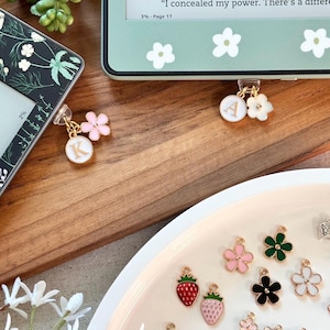 Może przedstawiać: Talerz z różnymi emaliowanymi zawieszkami, w tym kwiatami, truskawkami i sercem. Zawieszki są złocone i mają różne kolory, w tym różowy, czerwony, zielony, niebieski i czarny.