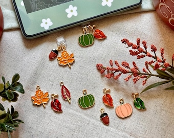 Könnte beinhalten: Ein E-Reader mit einer grünen floralen Hülle und dem Text "RACHEL GILLIG". Verstreut sind farbenfrohe Herbst-Charms, darunter Kürbisse, Blätter und Eicheln. Die Charms sind goldfarben mit Emaille-Details.